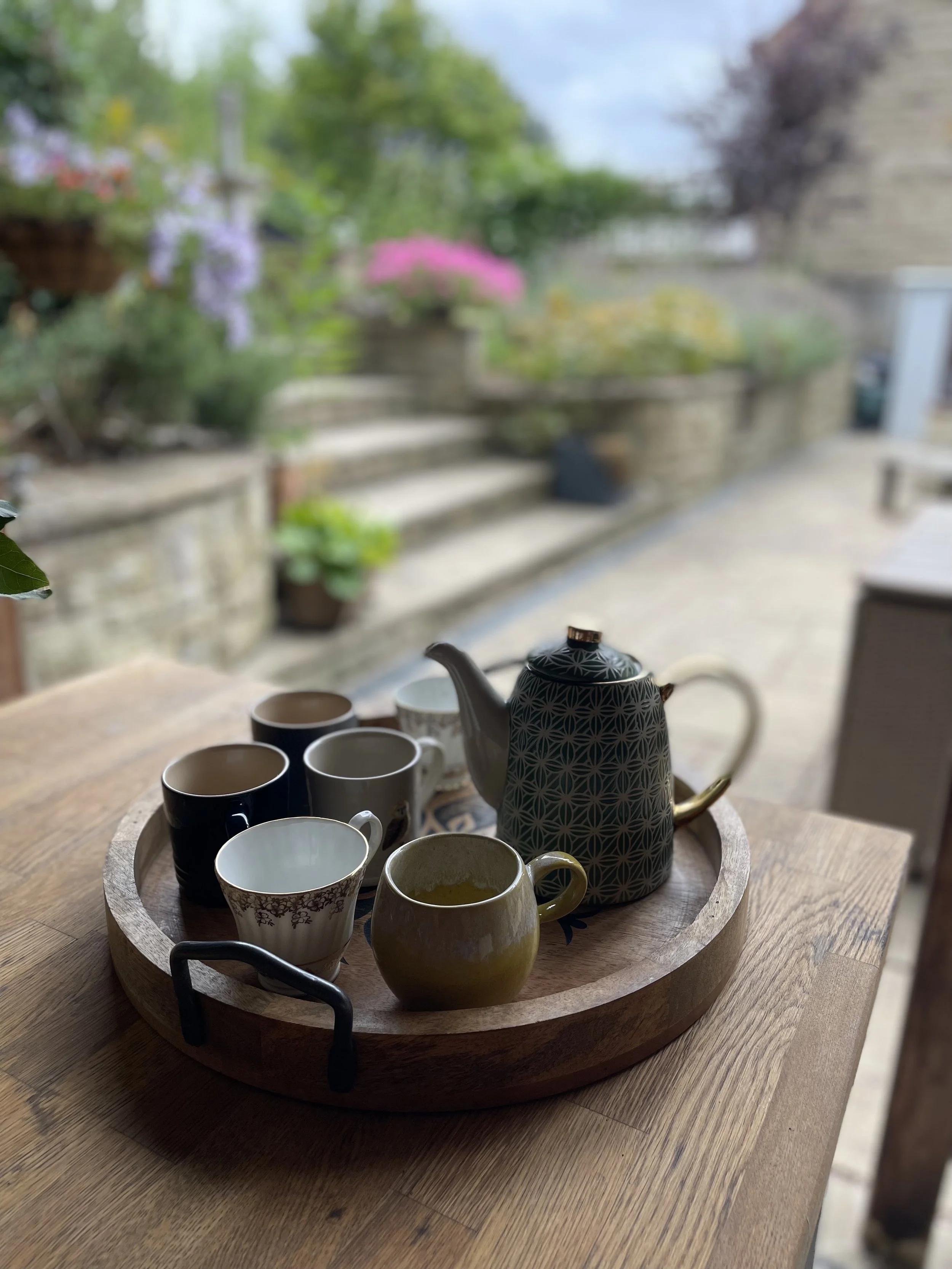 A wooden tray with a teapot and multiple teacups on a wooden table, with a blurred background of a garden with plants and stairs.