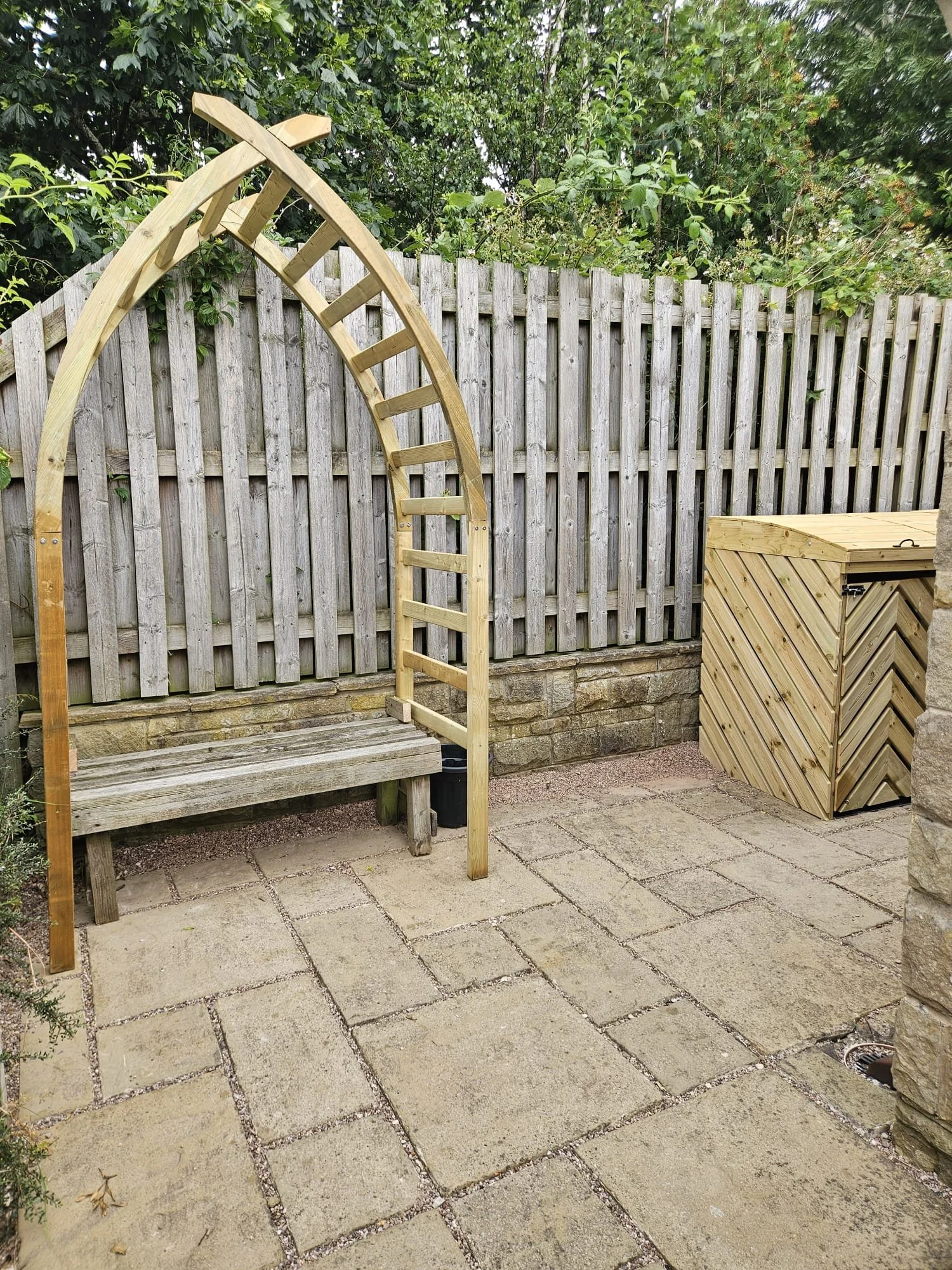 A small wooden arch with a ladder-like design stands next to a wooden bench on a tiled patio. A wooden storage box is nearby, and a weathered wooden fence runs across the background. Green trees and shrubs are visible behind the fence.