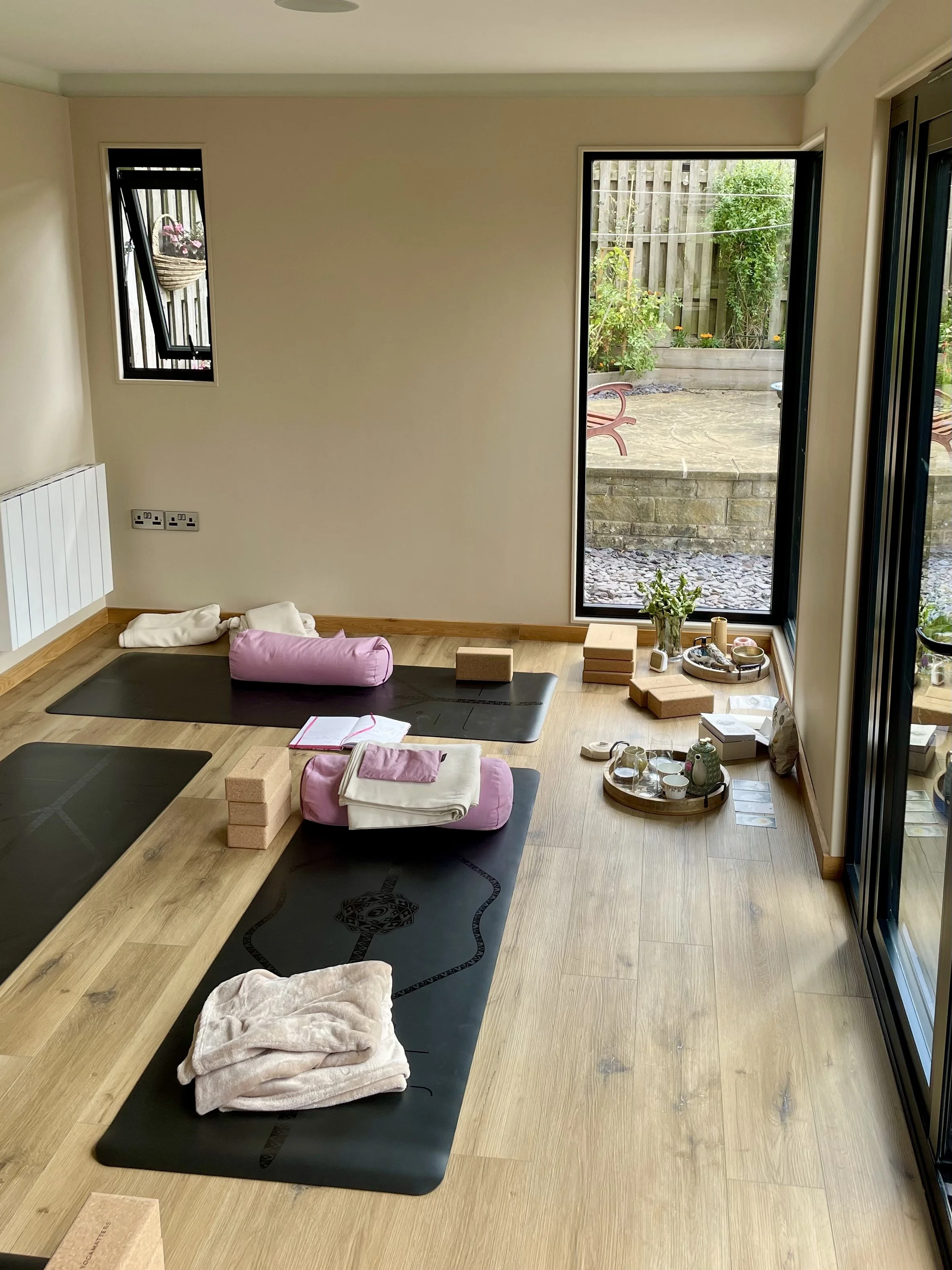 A yoga or meditation studio with black mats on a wooden floor, pink cushions, blankets, yoga blocks, a small white notebook, and various decorative items on a wooden tray. Large windows and doors reveal a patio with garden plants and wooden fencing.