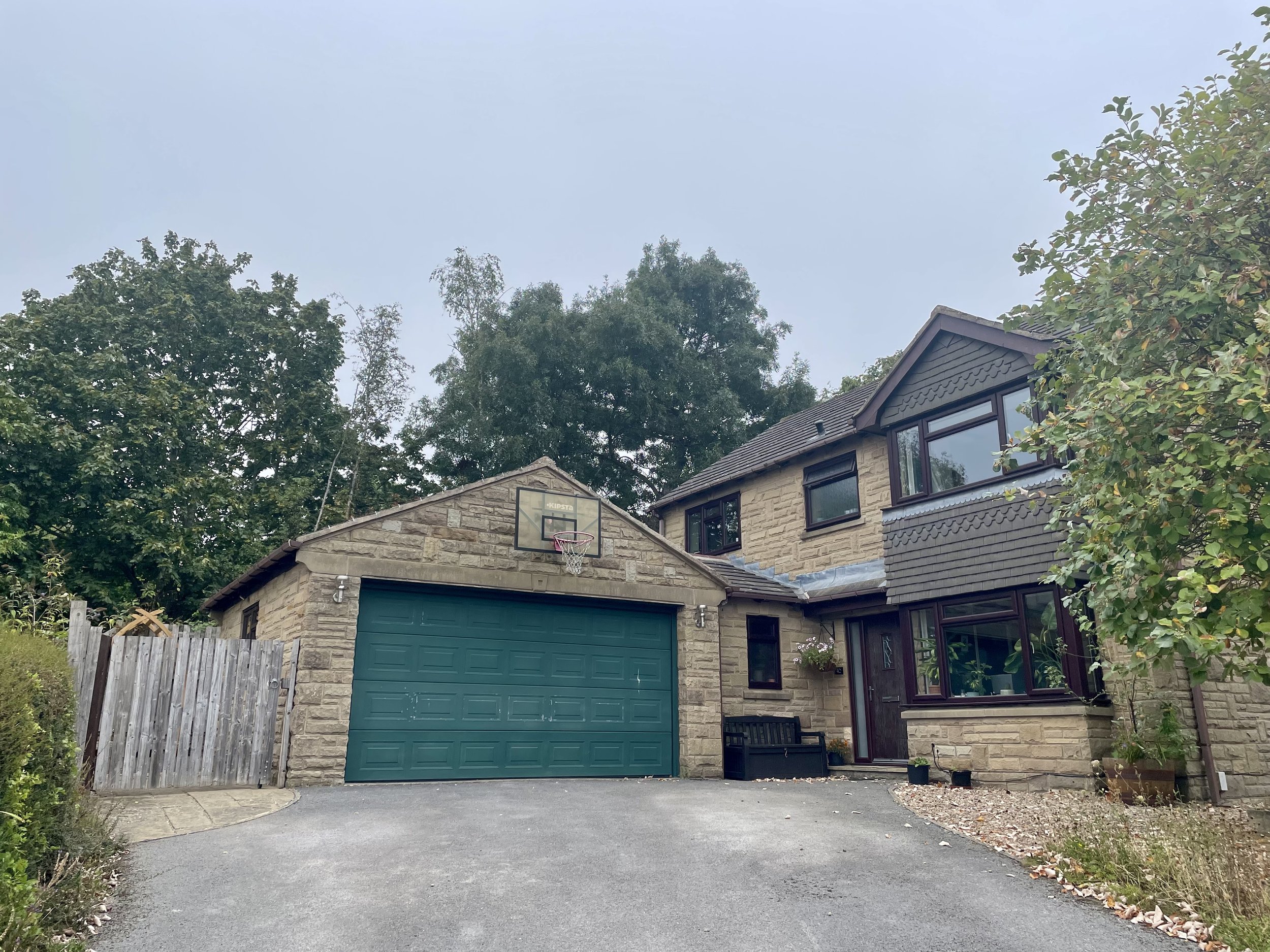 A residential house with a stone exterior, a green garage door, and black-framed windows. There is a basketball hoop mounted on the house over the garage, and a wooden fence on the left side. The driveway is paved, and there are trees and foliage sur