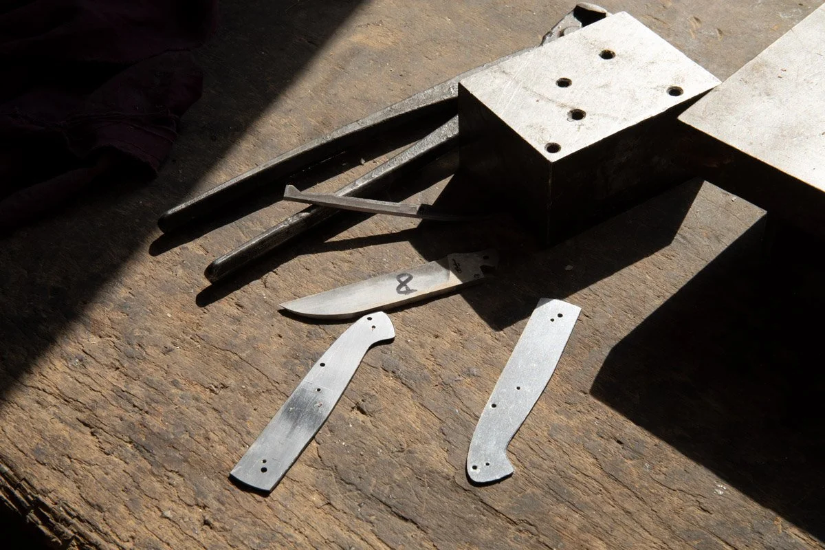 Outils de fabrication de couteaux, comprenant deux lames métalliques, deux pièces de fixation, un coin et une lime, posés sur une surface en bois, avec un moule rectangulaire en métal à côté.