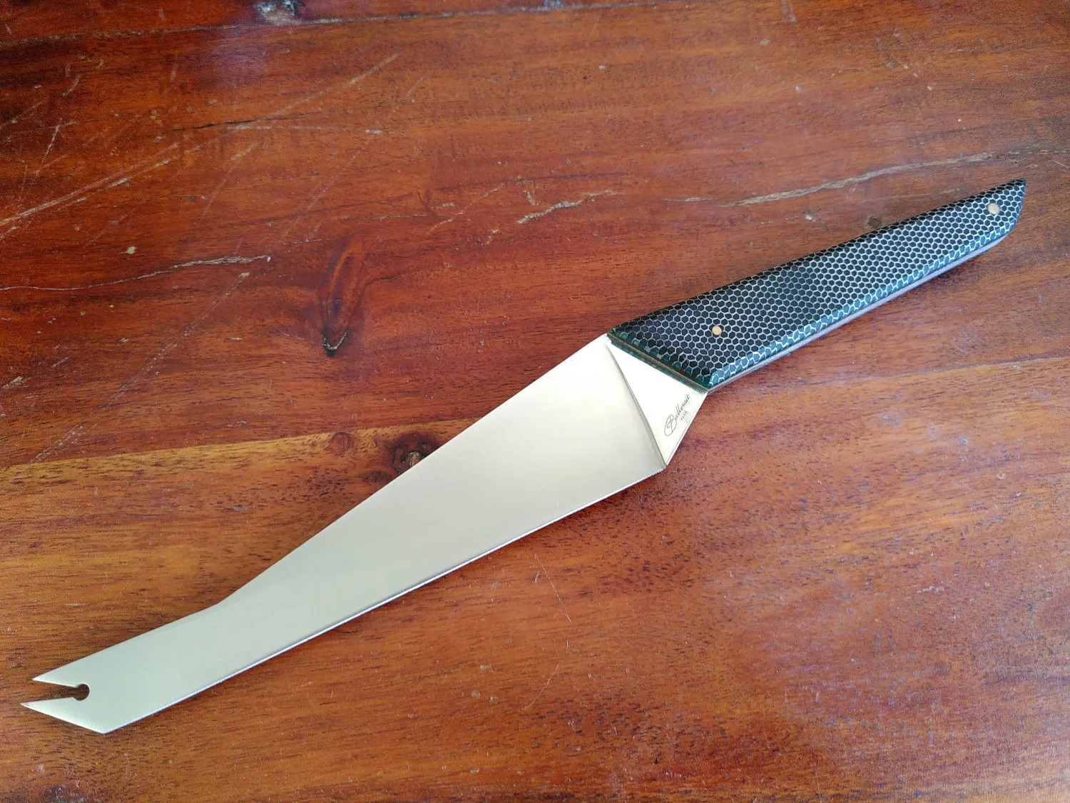 Un couteau à fromages CheeseSlayer, manche en nid d'abeille, posé sur une surface en bois.