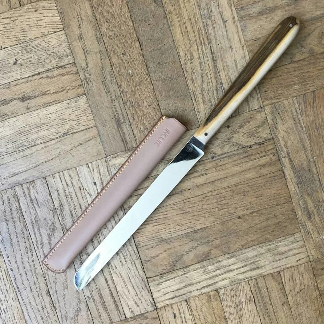 Couteau de métier pour doreur avec manche en bois de pistachier, posé sur un parquet en bois clair, avec un étui sur-mesure en cuir rose à côté.