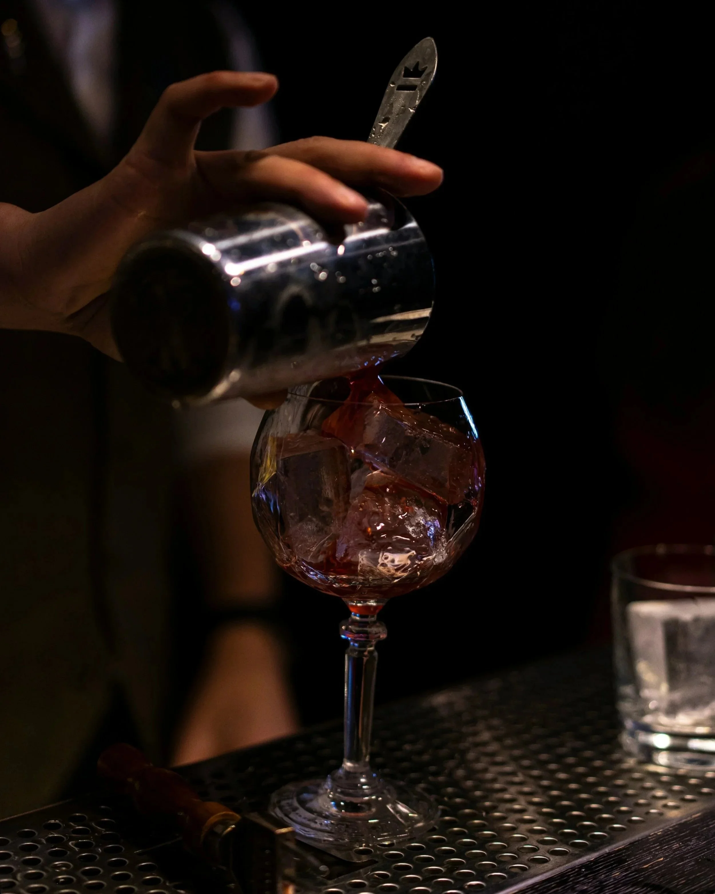 A hand pouring red liquid from a silver shaker into a wine glass filled with ice, set against a dark background.
