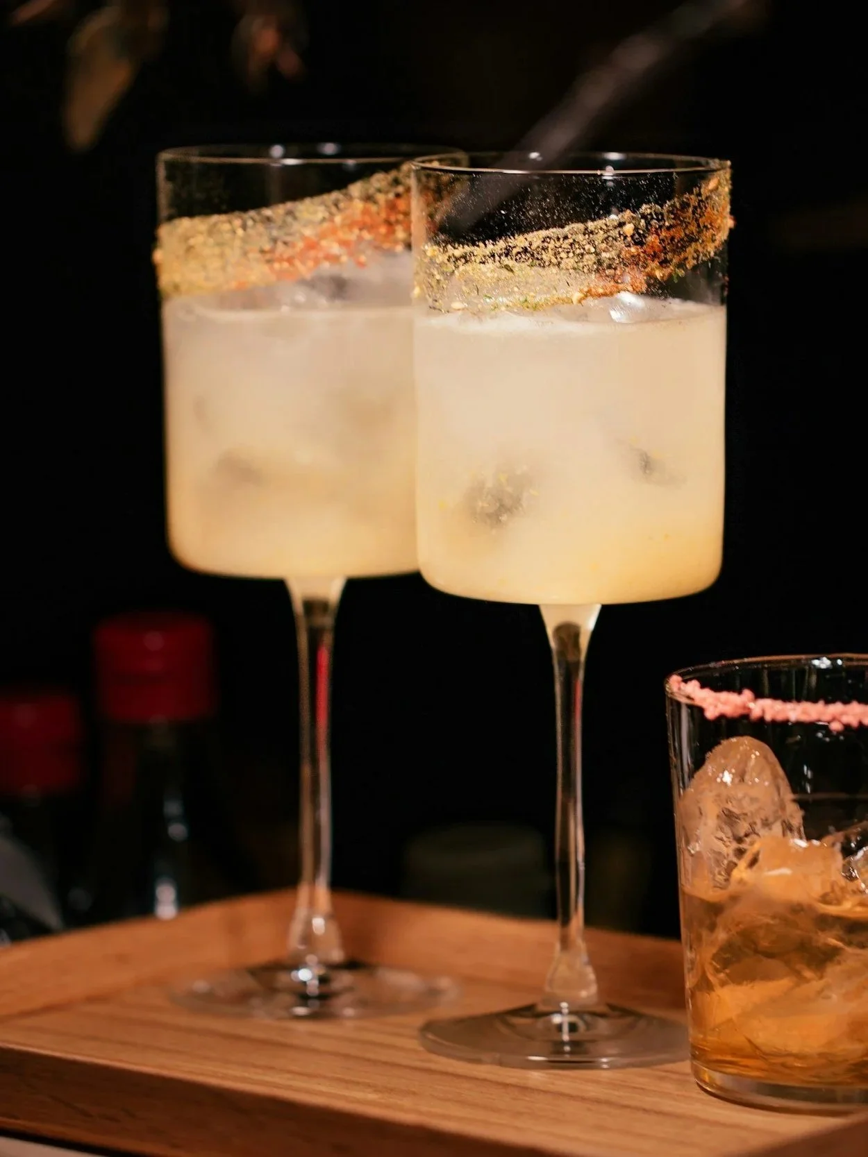 Two champagne glasses with salted rim and ice cubes inside, on a wooden table, with a cocktail glass containing ice and pink salt in the background.