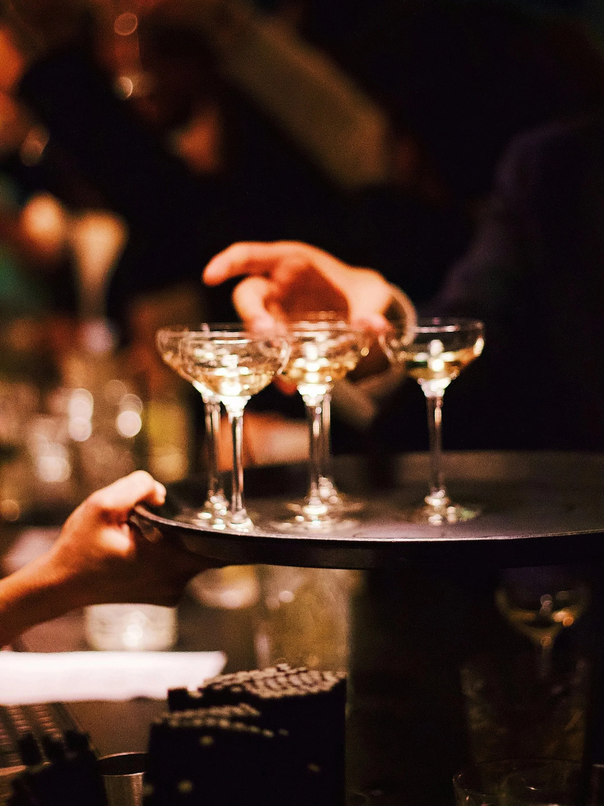 A person holding a tray with three cocktail glasses, each filled with a drink, in a dark setting with blurred lights in the background.