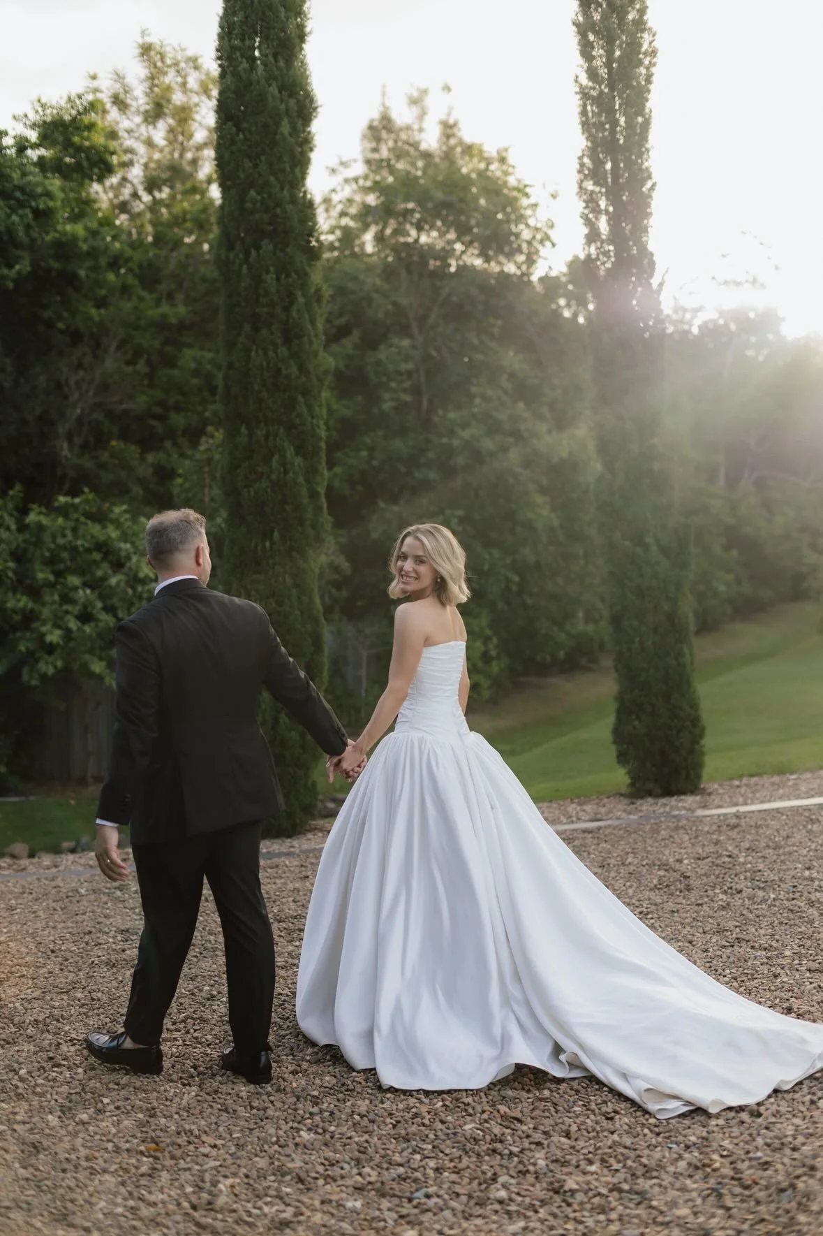 A newlywed couple holding hands outdoors during sunset, with the bride in a white strapless wedding gown and the groom in a black tuxedo, surrounded by tall green trees.