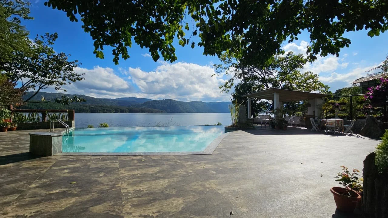 A swimming pool overlooking a lake with mountains in the background, surrounded by trees and outdoor furniture.