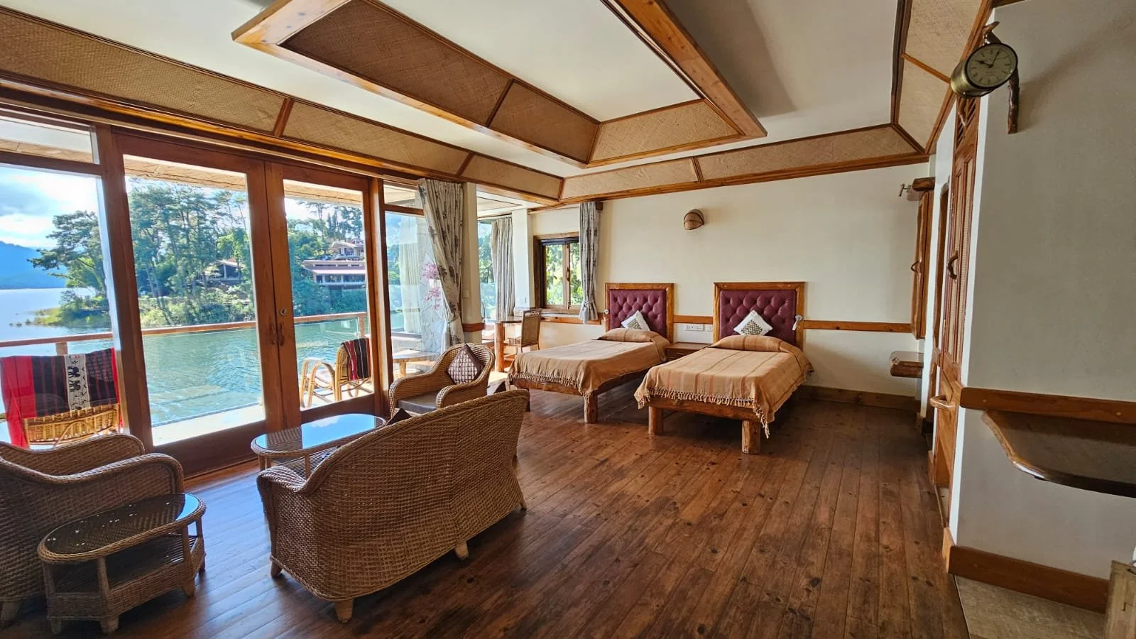 Living room with two single beds, wicker chairs, a table, large glass doors leading to a balcony with view of lake and trees, wooden floor and ceiling, clock on wall.
