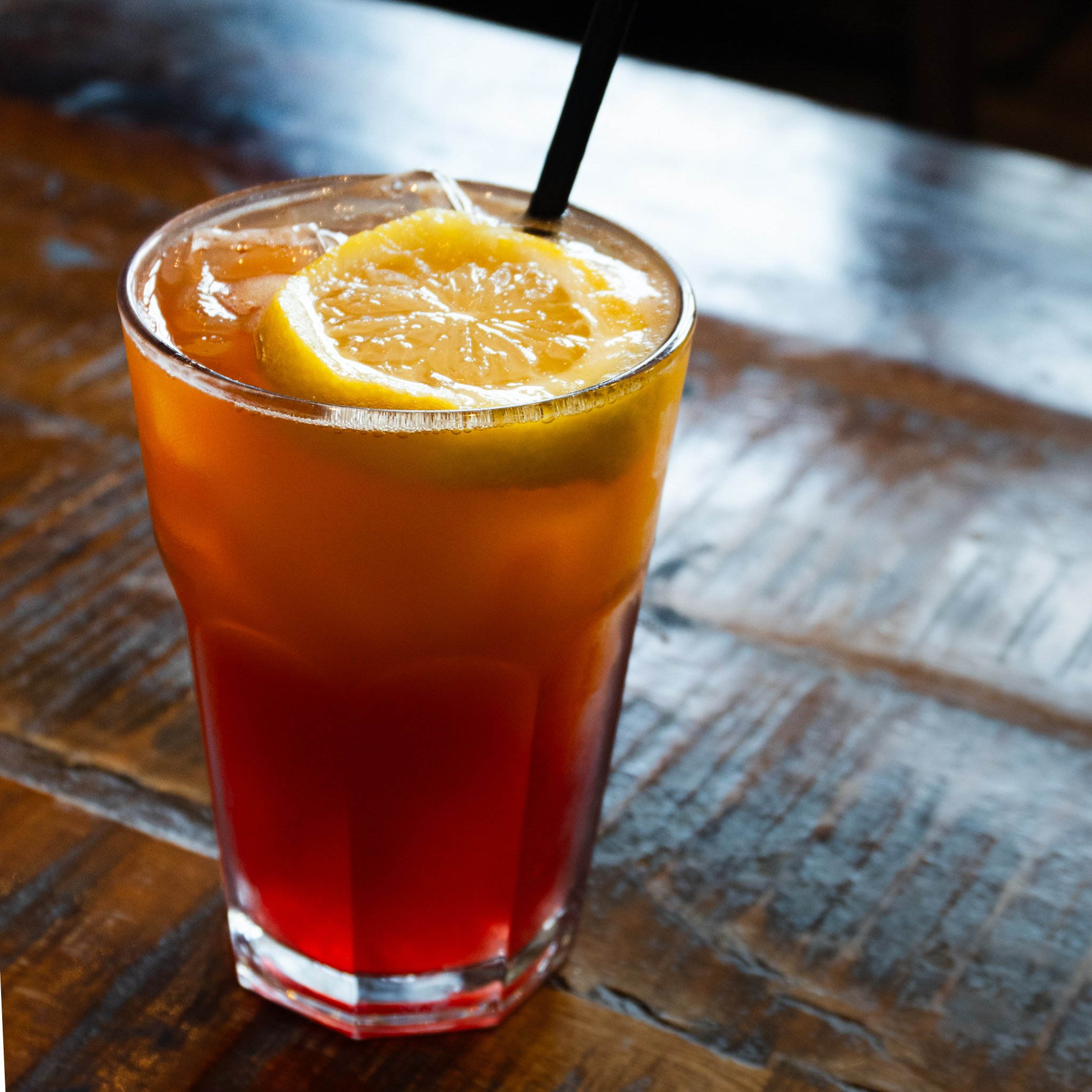 A glass of orange-colored beverage with ice cubes and a slice of lemon inside, placed on a wooden surface.