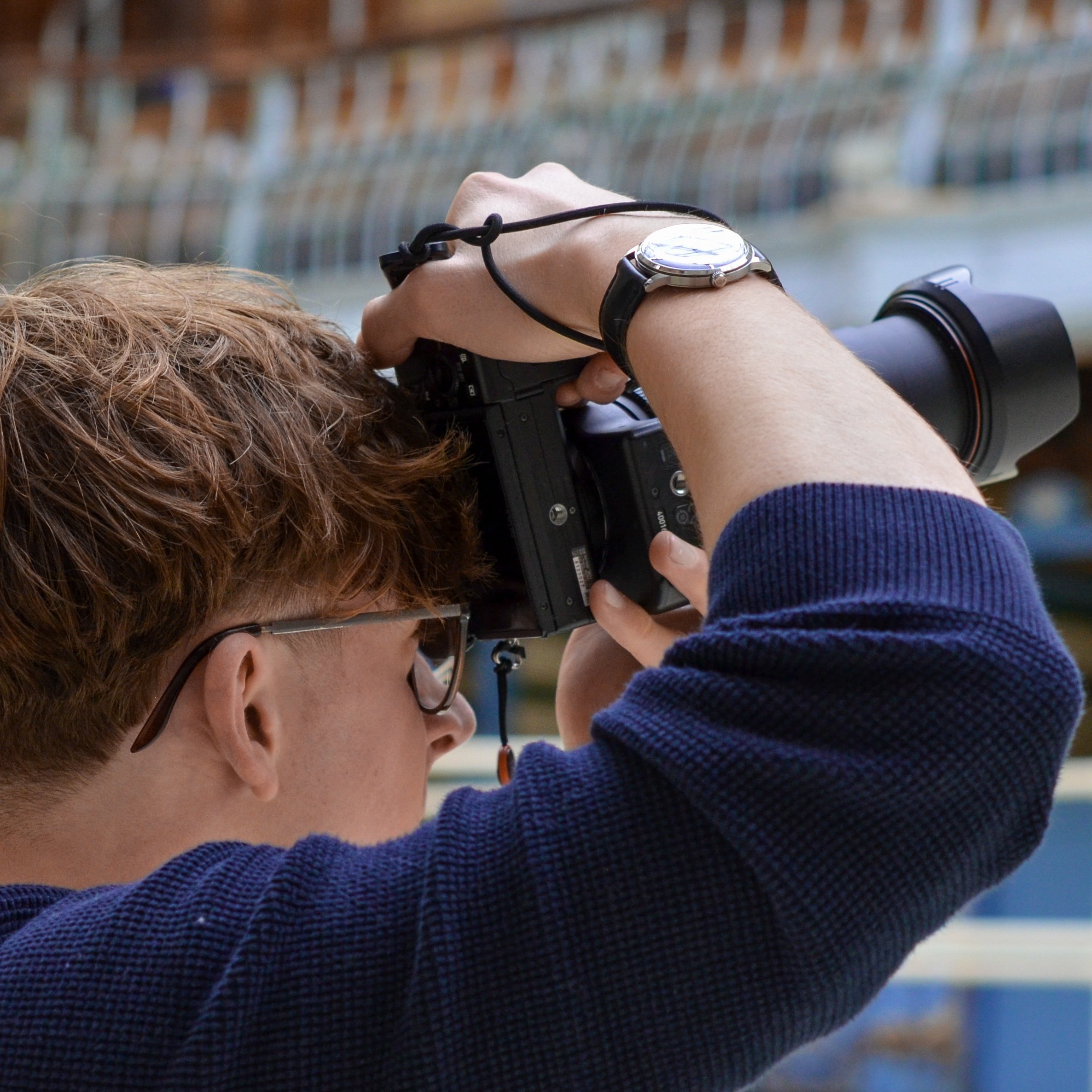 A person with reddish-brown hair wearing glasses is taking a photograph or looking through a camera, with their hand on the camera's focus ring, outdoors near a body of water with a blurred railing or fence in the background.