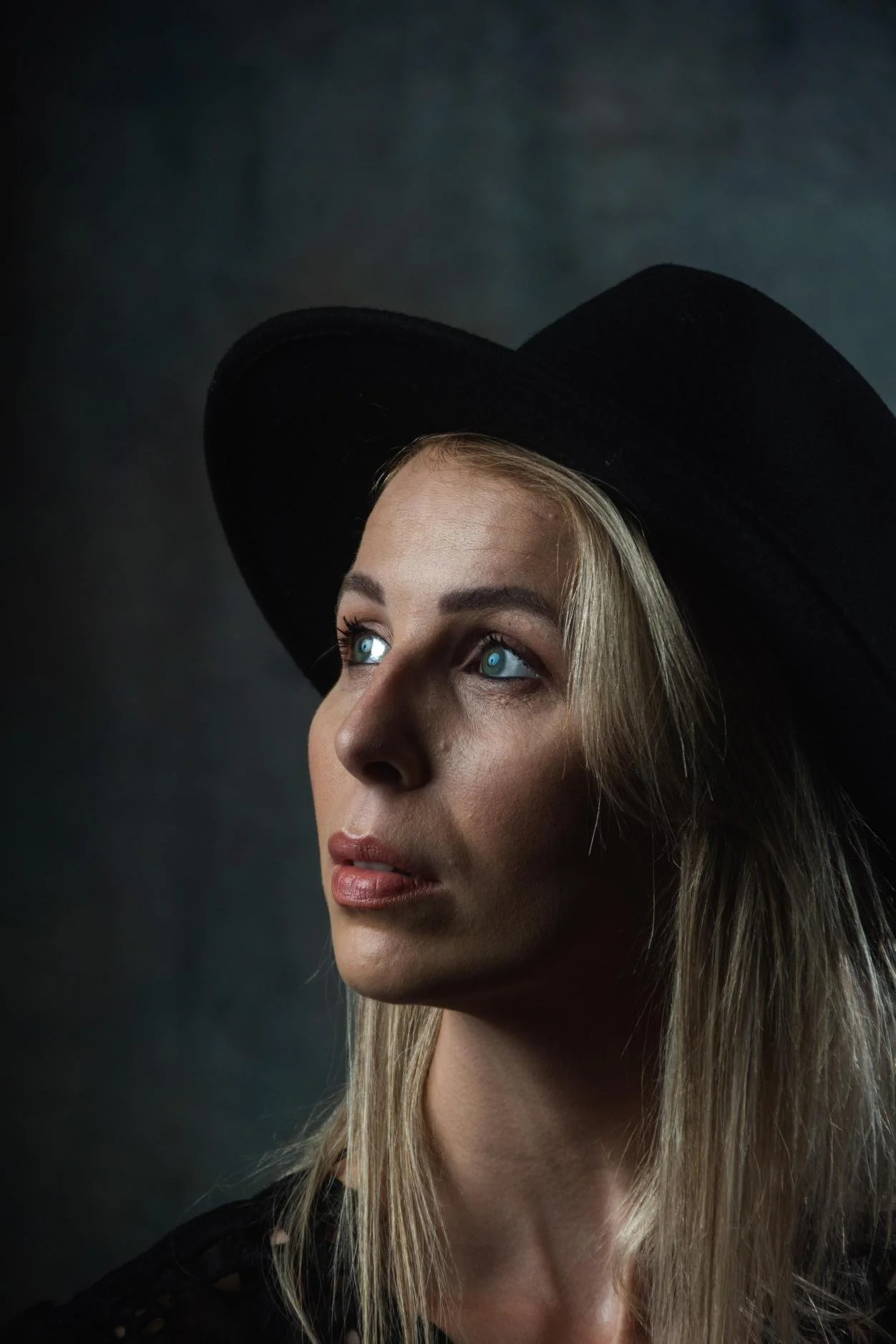 A woman with blonde hair and blue eyes wearing a wide-brimmed black hat, looking slightly upward against a dark background.
