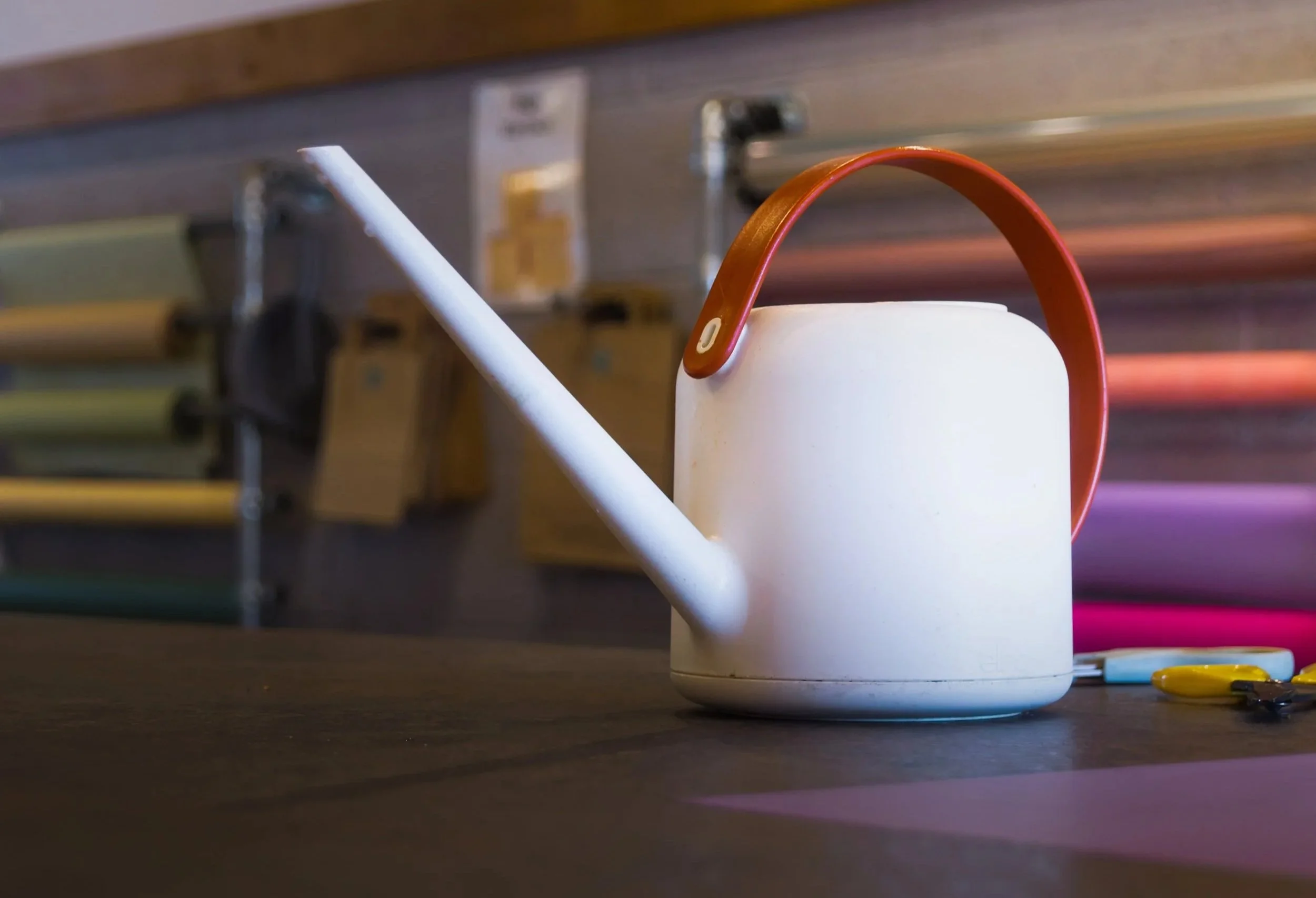 A white mug with a red handle on a dark surface, with colorful pieces scattered nearby, in front of a blurred background of shelves and objects.