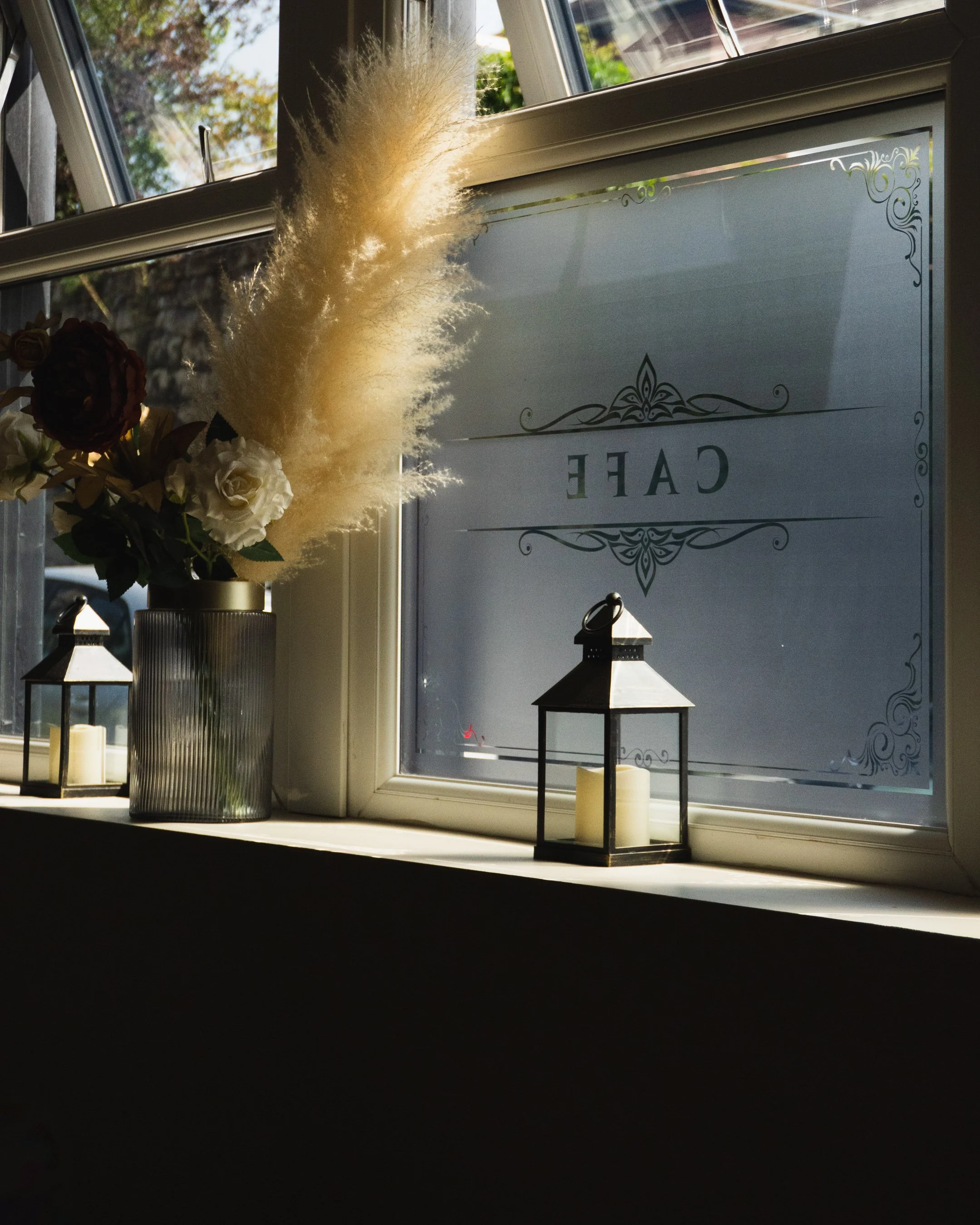 A window sill decorated with a tall gray vase holding white and dark red flowers, a potted plant with fluffy cream-colored pampas grass, two candle lanterns, and a sign with the word 'CAFE' on it.