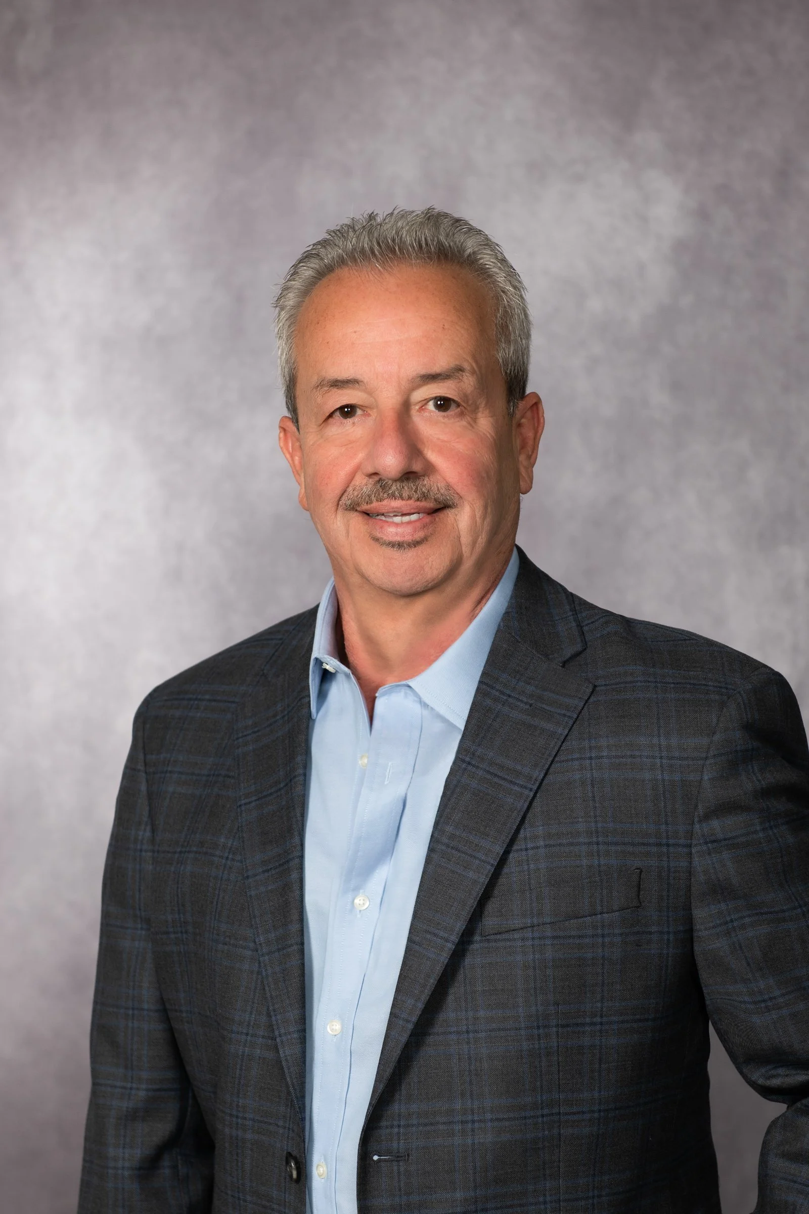 A professional headshot of a middle-aged man with gray hair, a mustache, and a slight smile, dressed in a dark checkered suit jacket and light blue collared shirt, standing against a neutral gray background.