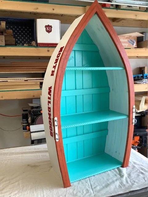 A boat-shaped wooden shelf painted turquoise inside with two shelves, with red trim and white sides, in a workshop setting.