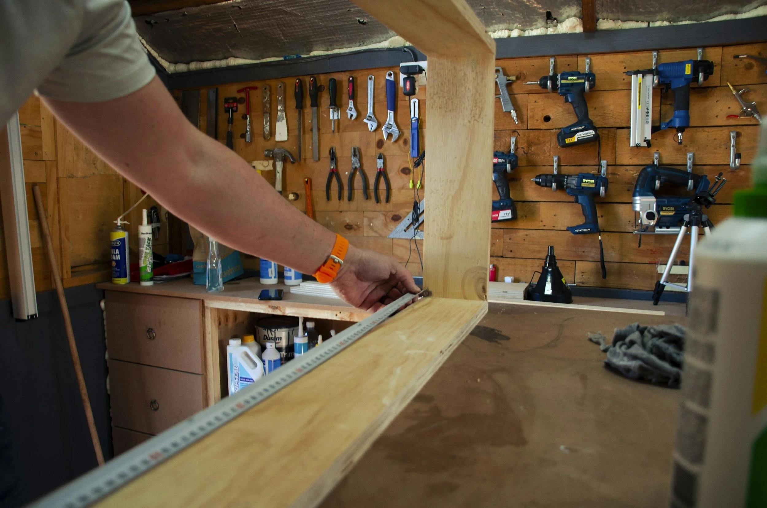 A person working on a woodworking project in a workshop with tools hanging on a wooden wall, including screwdrivers, pliers, and power drills, as well as bottles of glue and other supplies on a workbench.
