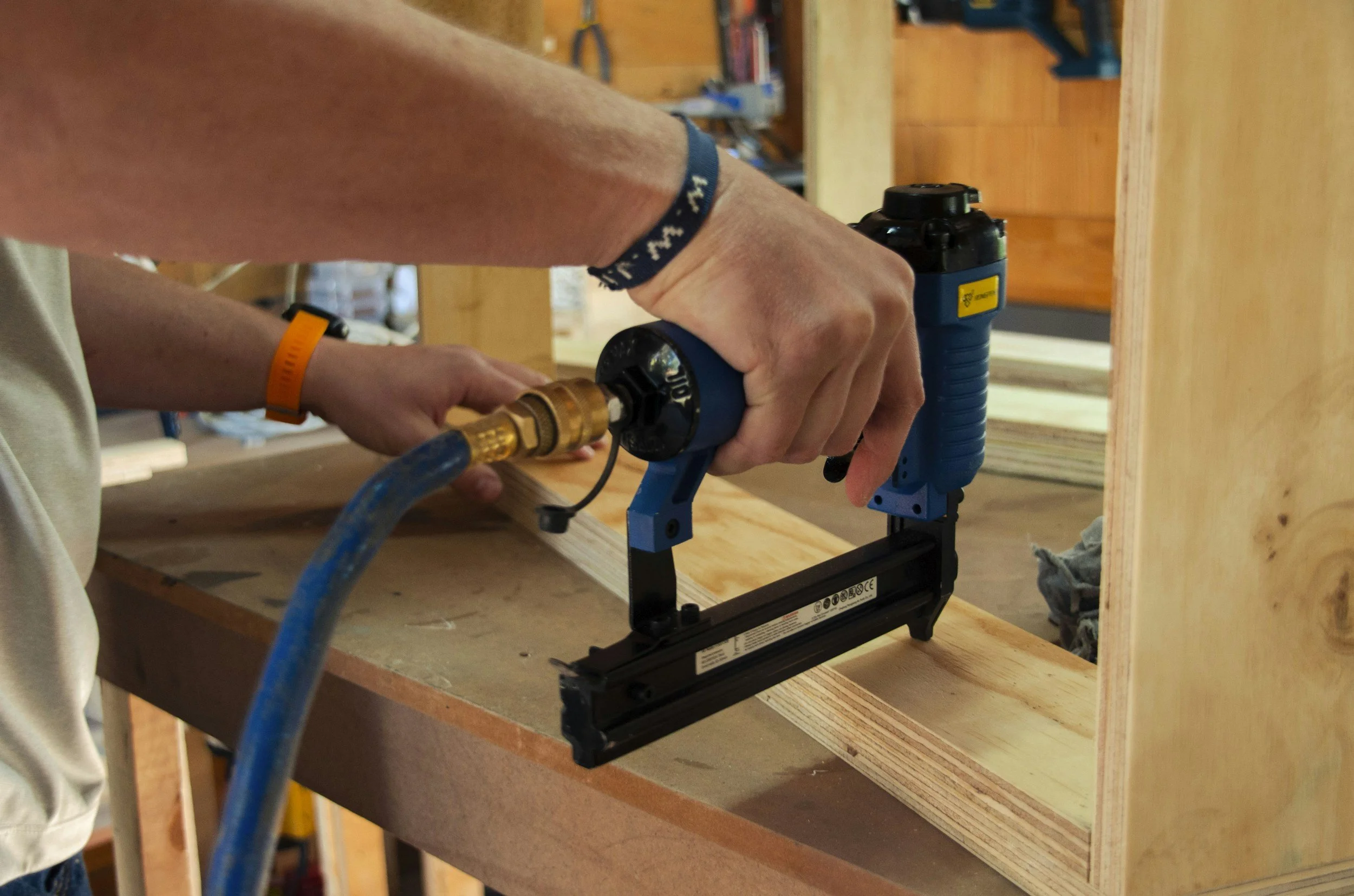Person using a pneumatic nail gun on a piece of wood in a woodworking shop.
