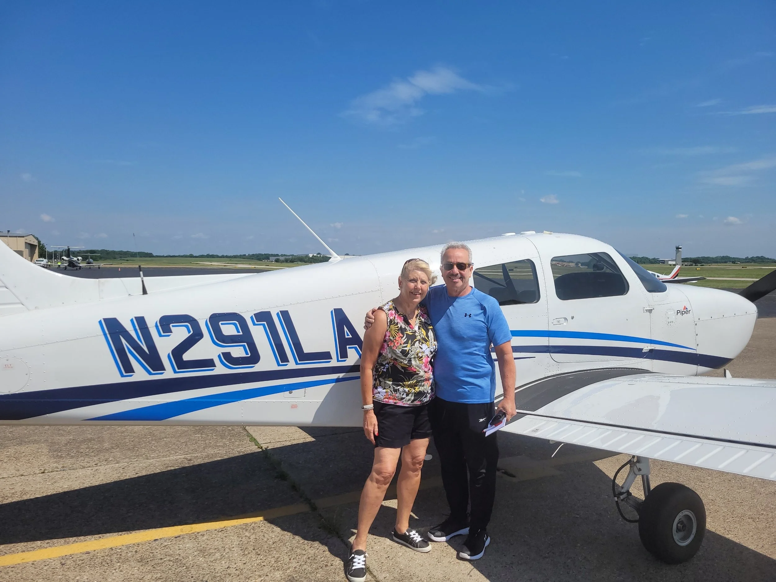 A smiling elderly woman and a man stand in front of a white small airplane on a sunny day at an airport. The woman is wearing a floral top and black shorts, and the man is wearing a blue t-shirt and black shorts. They are holding a clipboard and standing close together.