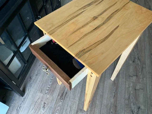 Wooden table with an open drawer, showing miscellaneous items inside, located on a hardwood floor.