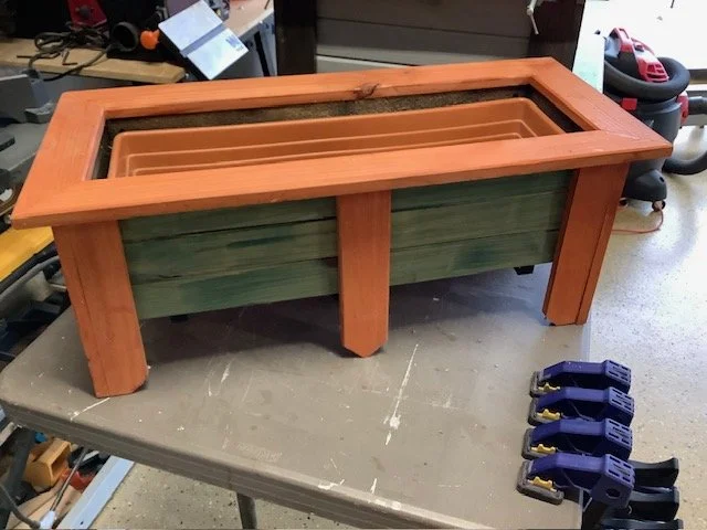 A wooden planter box with a rectangular glass top, placed on a workbench in a garage or workshop. There are clamps and various tools nearby.
