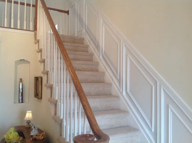 Indoor staircase with beige carpet, white banisters with wood handrail, white paneled wall, and a small side table with a lamp and decorative items.