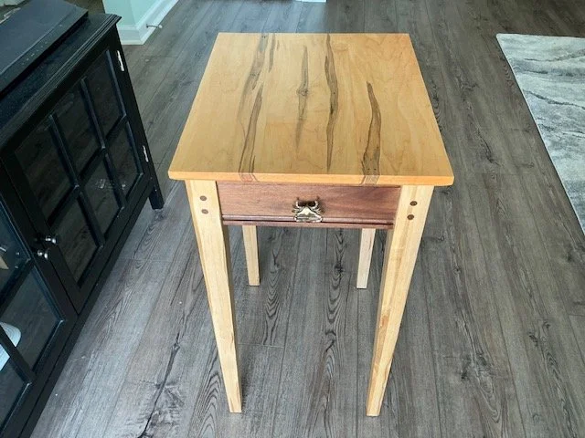 Wooden side table with a single drawer and curved metal handle, placed on a dark wood floor next to a black cabinet.