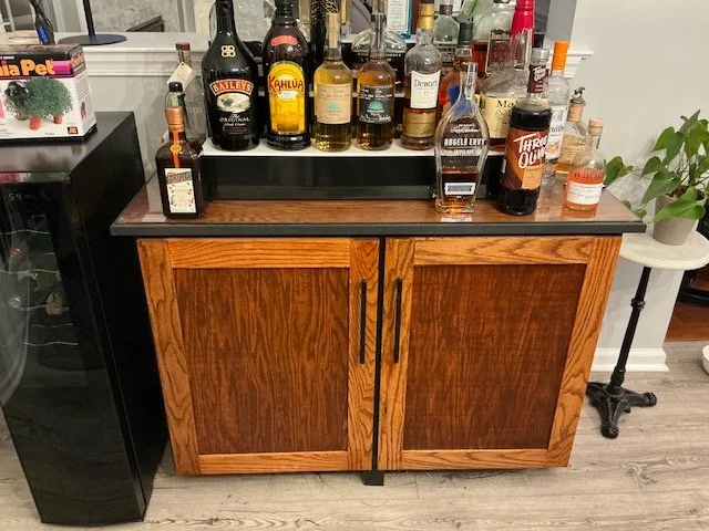 A wooden cabinet with two doors and black handles, topped with a black countertop. Various bottles of alcohol, including whiskey, tequila, and liqueurs, are arranged on top. To the left is a black mini fridge, and to the right, there is a small round table with a potted green plant.