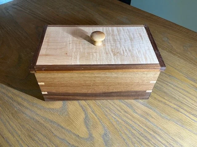 A wooden storage box with a small round handle on the lid, sitting on a wooden table.