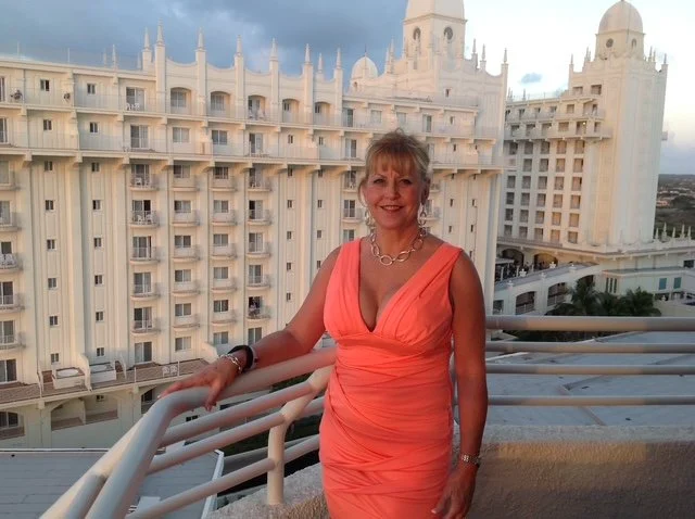 A woman in an orange dress standing on a balcony with a large white hotel building in the background.