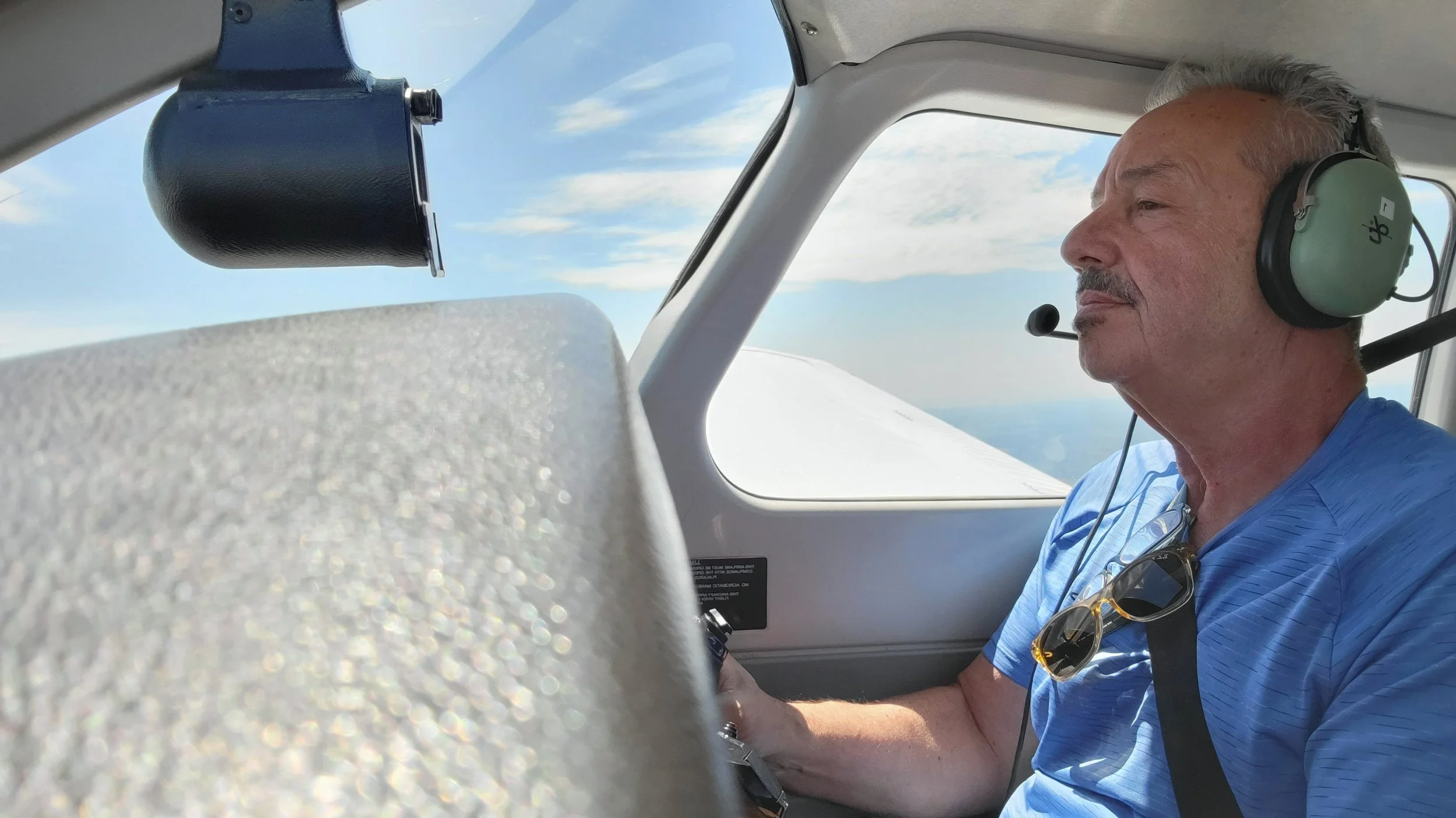 A man with gray hair wearing a blue shirt and sunglasses hanging on his shirt sits in the cockpit of a small airplane, wearing green noise-canceling headphones, with the sky and clouds visible through the window.