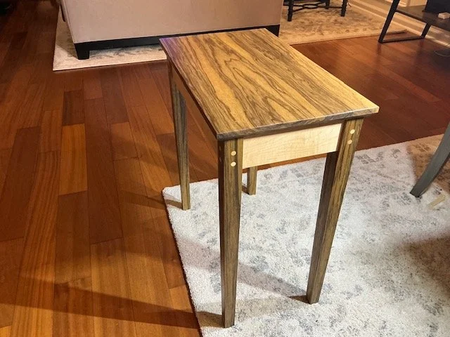 A small wooden side table with a rectangular top, standing on four tapered legs in a room with hardwood floors and a gray and white area rug.