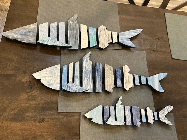 Three wooden fish-shaped art pieces painted in shades of blue, gray, and white, arranged on a wooden table.