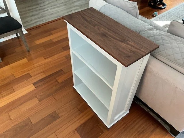 A small white bookshelf with three shelves and a dark brown wooden top, situated next to a light-colored sofa on a wood floor.