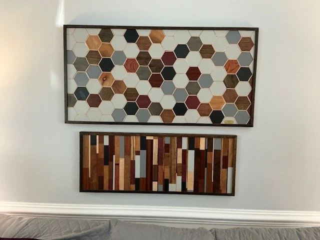 Two framed wood art pieces hanging on a white wall above a gray sofa. The top piece features a honeycomb pattern with hexagonal wooden tiles in various shades of brown, gray, black, and cream. The bottom piece consists of vertical rectangular wooden slats in different colors and wood tones.