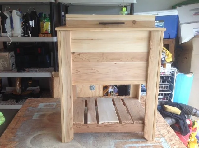 Unfinished wooden nightstand or small table with four legs and an open bottom shelf, placed on a workbench in a garage or workshop.