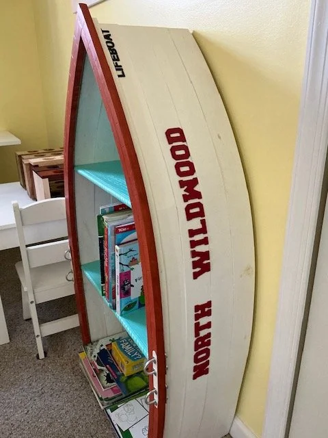 A bookshelf designed like a boat, labeled 'North Wildwood' and 'Lifeguard,' with colored shelves holding children's books, positioned next to a white chair in a room with yellow walls.