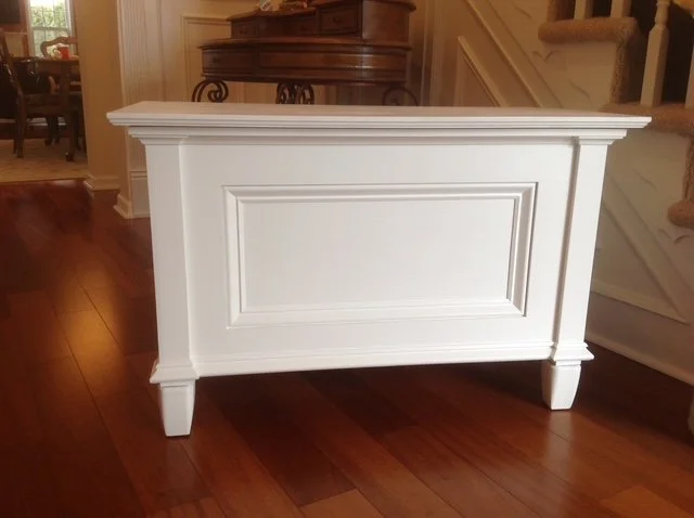 A white wooden storage chest with panel details, placed on a hardwood floor in a home interior.