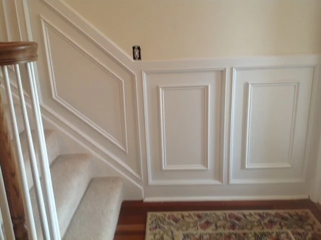 Interior view of a staircase with carpeted steps and a white decorative wall paneling, with a small section of a floral area rug visible on the hardwood floor.