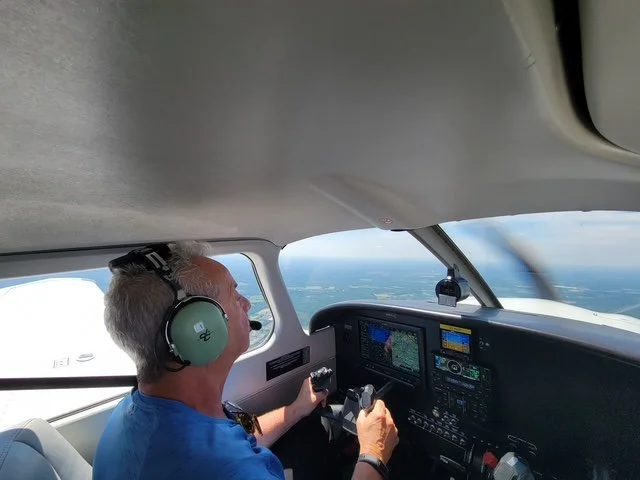 A man flying a small airplane, wearing headphones and a blue shirt, inside the cockpit with instrument panels and a view of the sky and clouds outside.