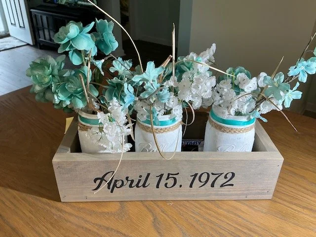 Wooden box with four Mason jars filled with decorative blue and white flowers, and the date April 15, 1972 written on the front.