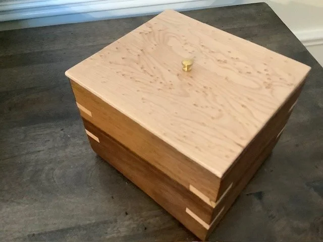 A wooden jewelry box with a small brass knob on the lid, sitting on a dark wooden surface.