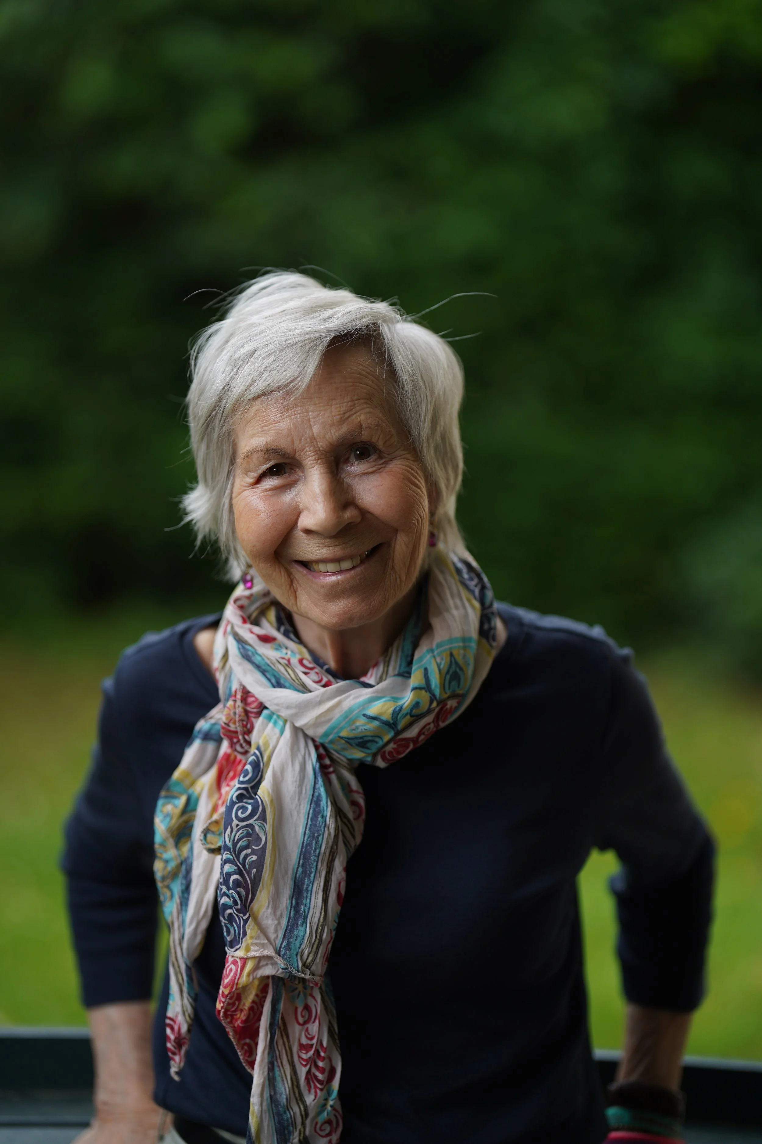 Eine alte Frau mit kurzen weißen Haaren, lächelnd, trägt eine bunte Schals, vor einem grünen, unscharfen Hintergrund.
