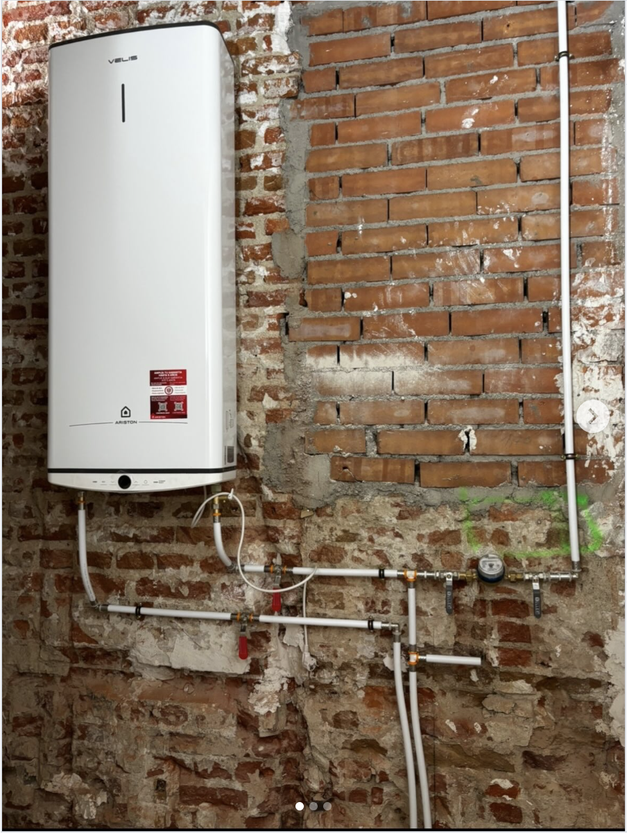 Calentador de agua instalado en una pared de ladrillos, con tuberías y un medidor de agua visible.