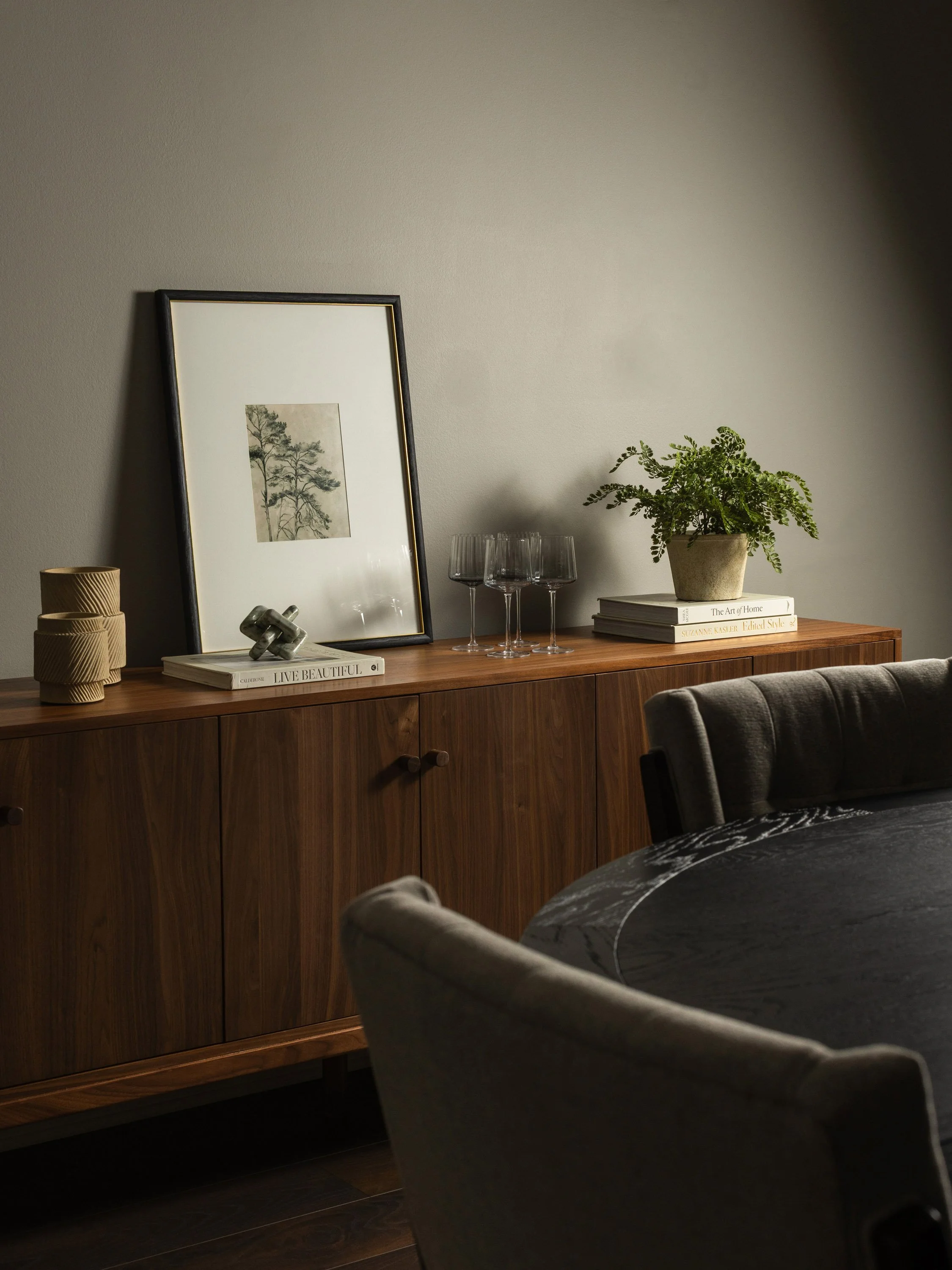 A wooden sideboard with framed artwork, decorative vases, a few books, a potted plant, three empty wine glasses, and a small metallic sculpture in a room with beige walls and dark flooring.