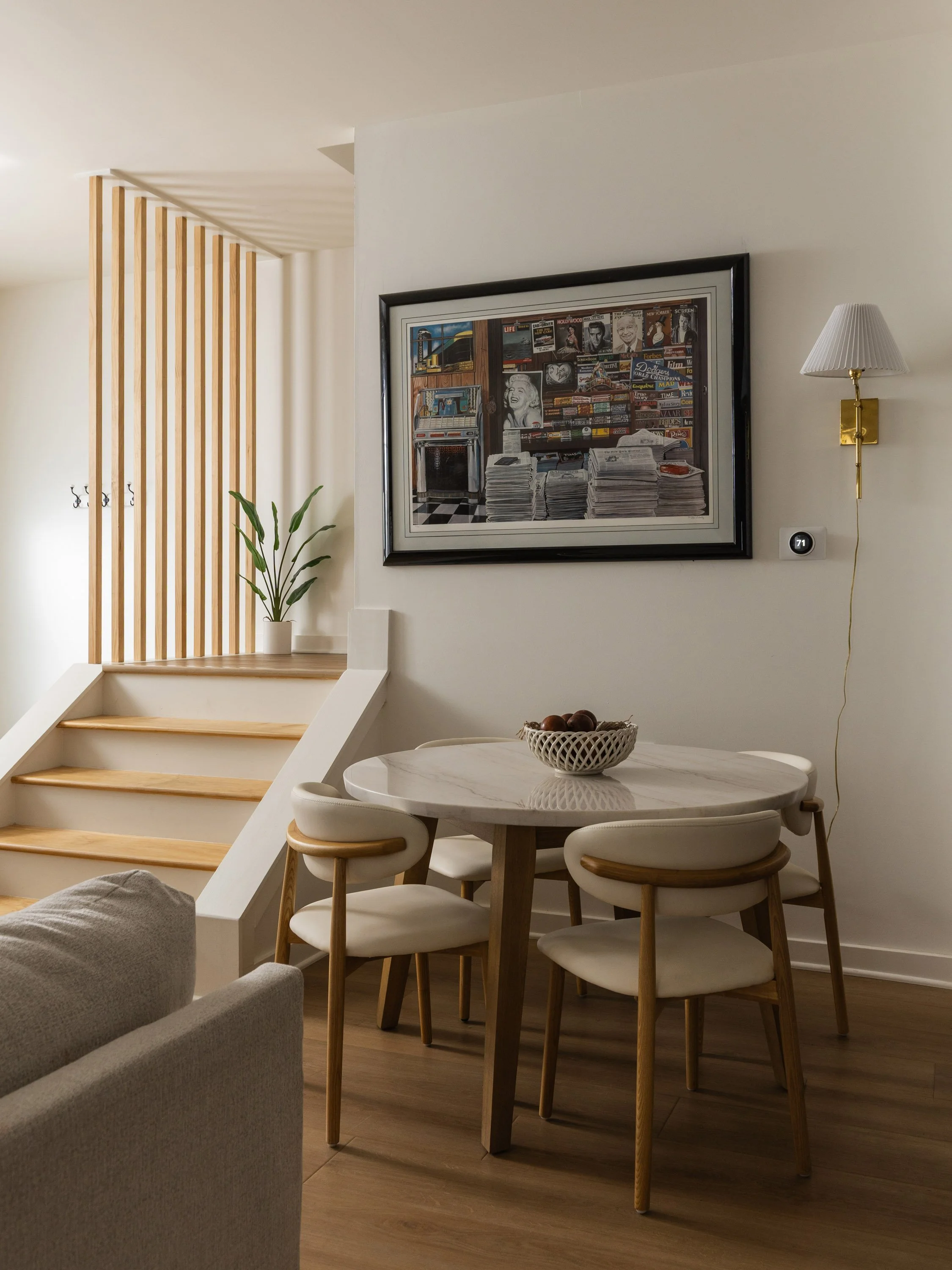 A cozy dining area with a round white table surrounded by four white chairs with wooden legs. There is a bowl of apples on the table. The background includes a wall with framed artwork, a wall-mounted lamp, and a staircase with wooden steps and a ver