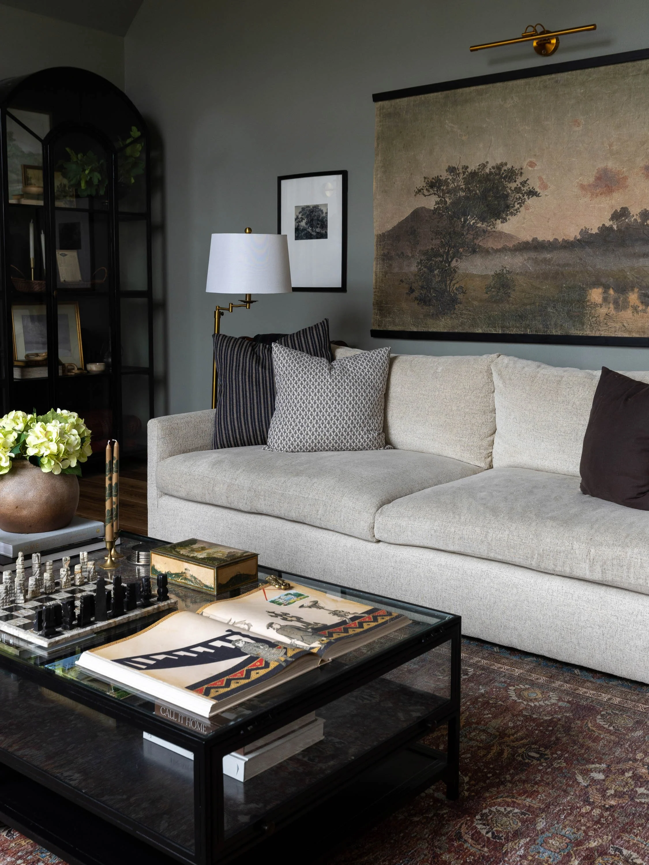 Living room with beige sofa, patterned pillows, wall art, a lamp, a black coffee table with books and a chessboard, a glass top with magazines, and a patterned rug.
