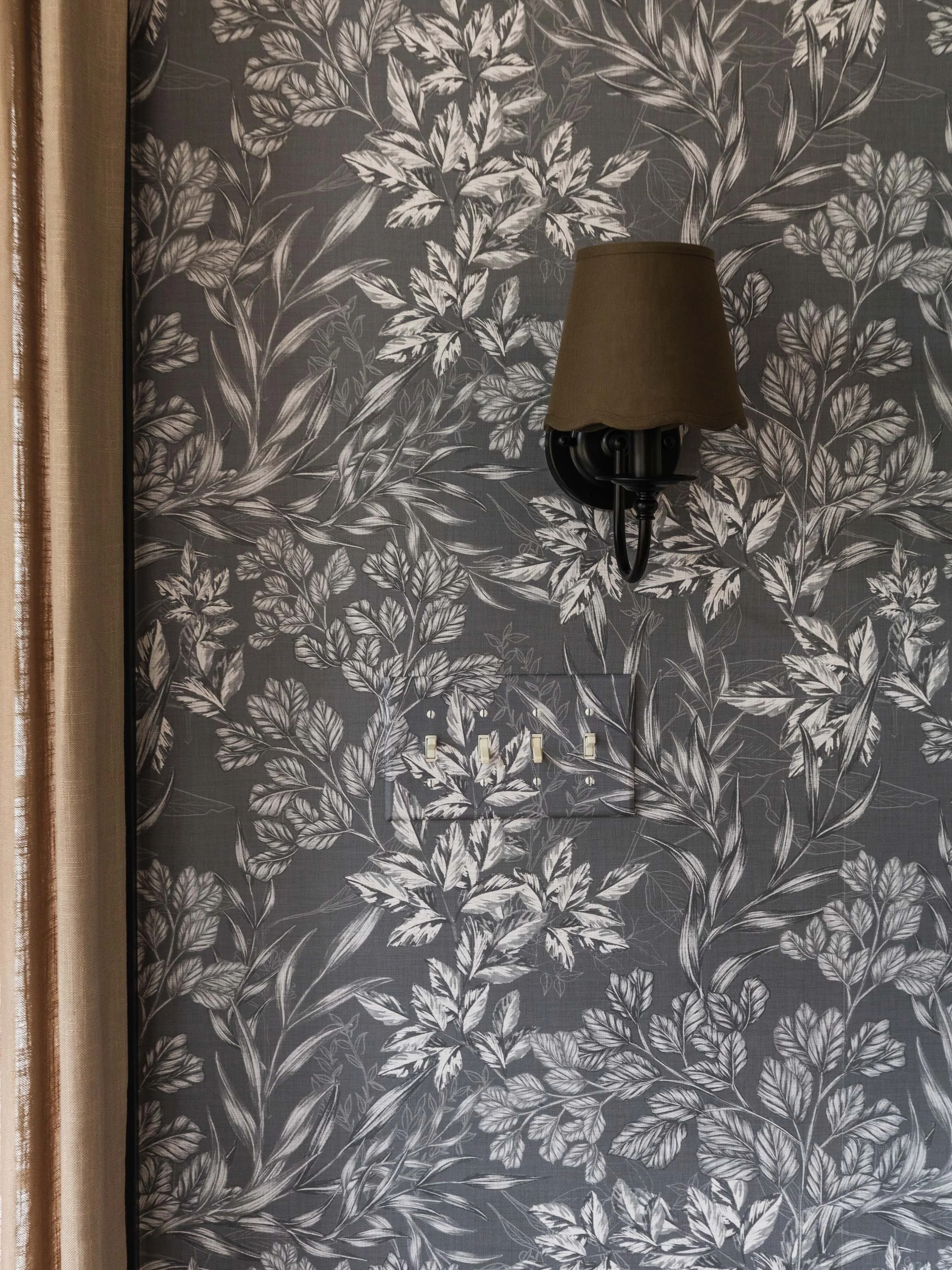 A wall with dark and light botanical wallpaper featuring various leaves and plants, with a wall-mounted lamp and a row of hooks below.