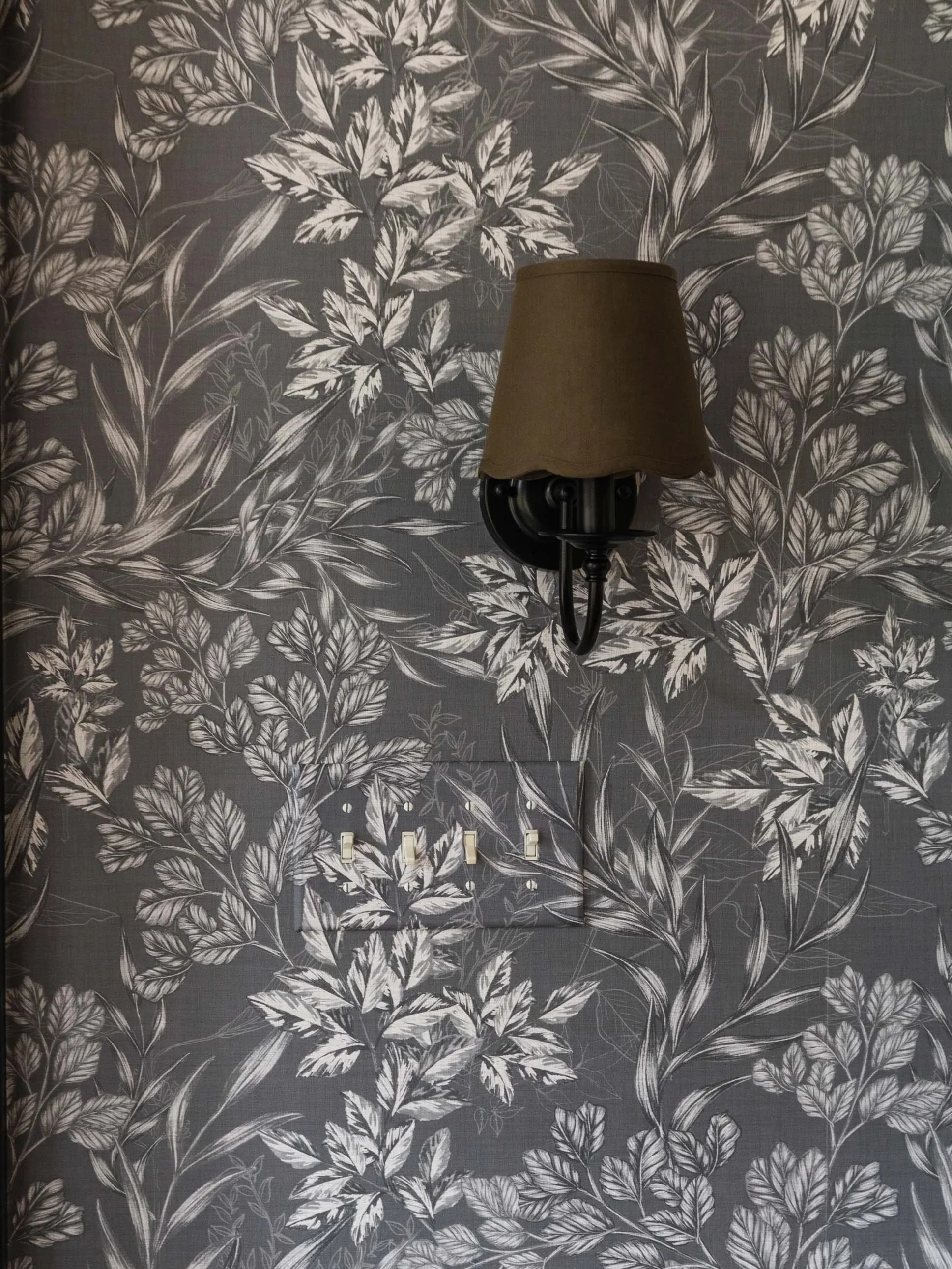 A wall with wallpaper featuring a floral pattern of white leaves and branches on a dark background, with a wall-mounted lamp and electrical outlets beneath it.