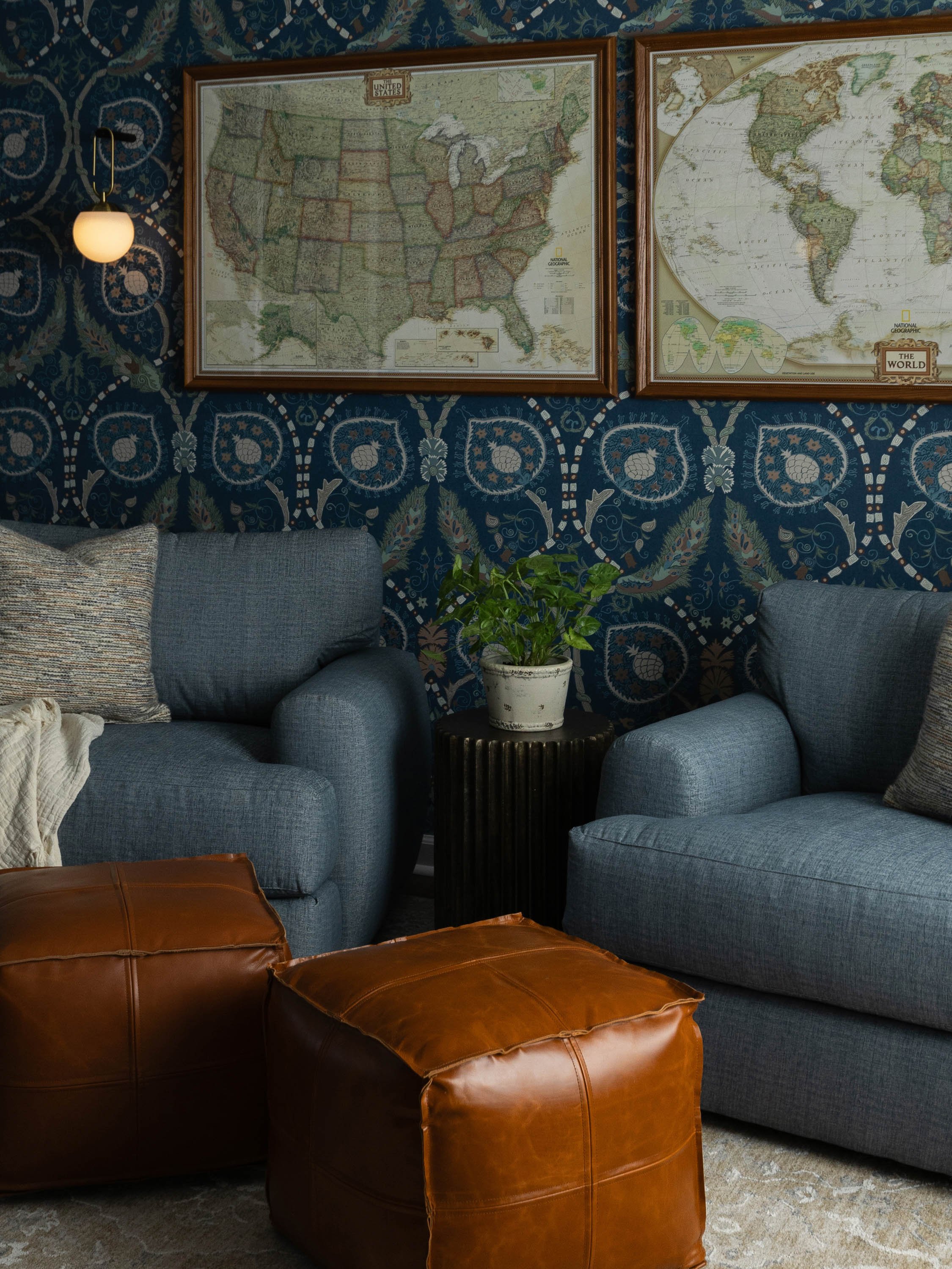 Living room with blue patterned wallpaper, two framed maps on the wall, a gray sofa with pillows, a small black side table with a potted plant, and a brown leather ottoman in the foreground.